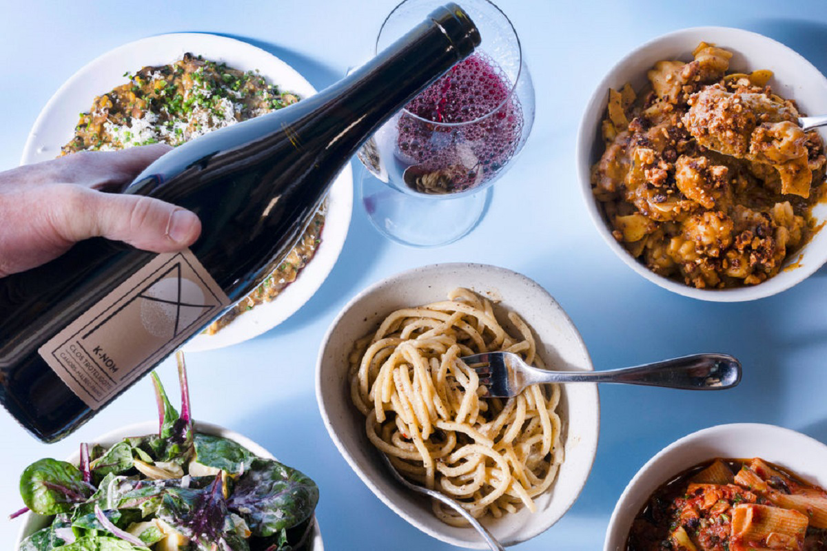 A hand pours red wine beside bowls of pasta, salad, and hearty vegetable dishes on a light blue table at Somebody People in Denver.
