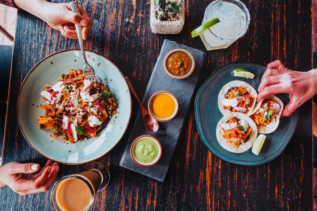 Cowtown-2 - DiningOut Tamayo tacos served with lime wedges and a side of salsa trio, paired with a plate of chilaquiles topped with crema, radish, and shredded meat on a rustic table.