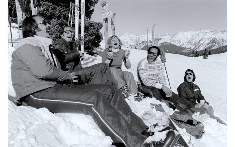A vintage black-and-white photo of a group of people sitting in the snow mid-mountain at Vail in the 1970s
