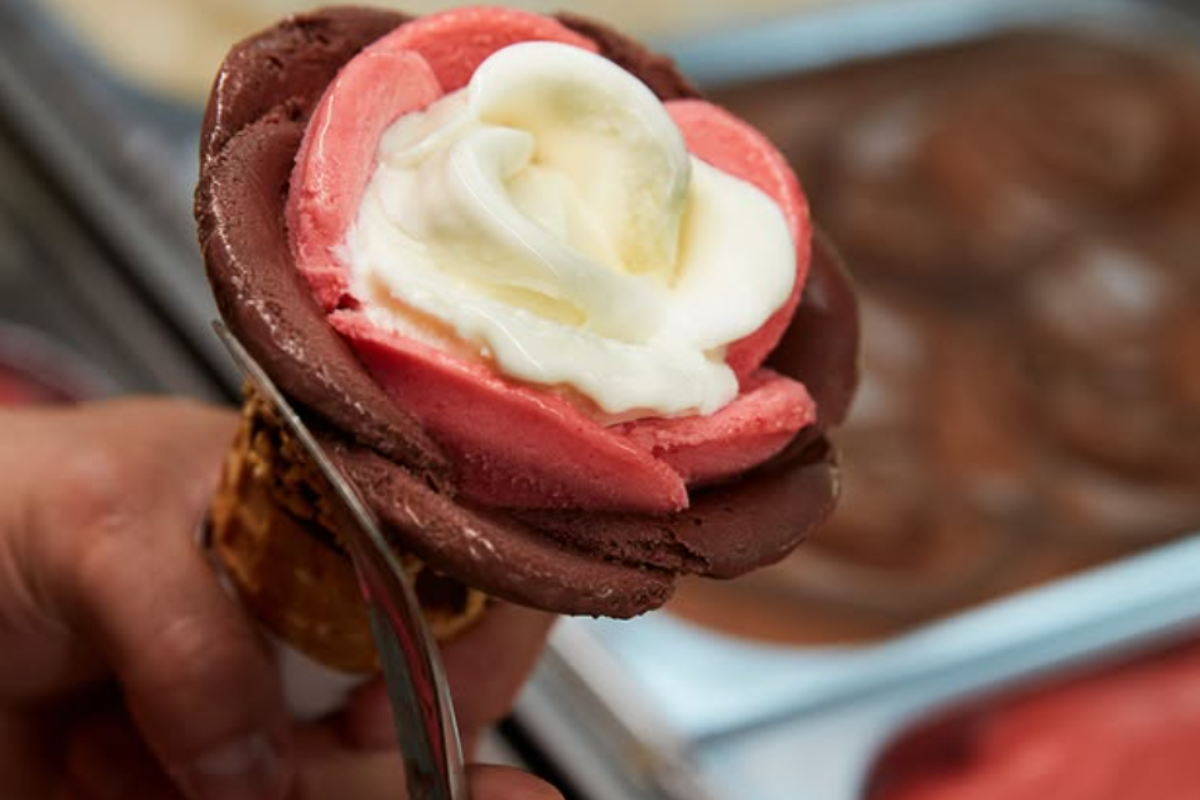 Close-up of a rose-shaped Amorino gelato cone with chocolate, strawberry, and vanilla gelato petals, being crafted with a spatula by hand.