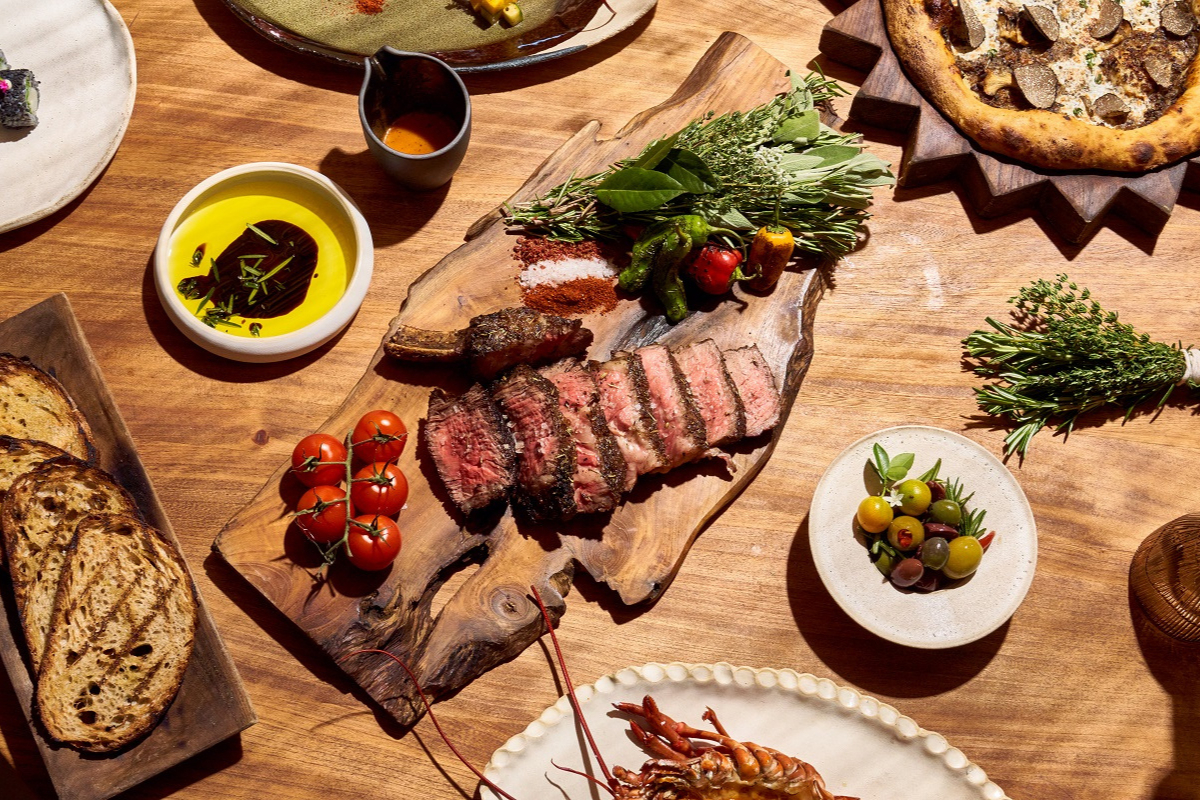 Grilled steak sliced on a wooden board with cherry tomatoes, herbs, grilled bread, olive oil, roasted peppers, and pizza on a rustic wooden table.

