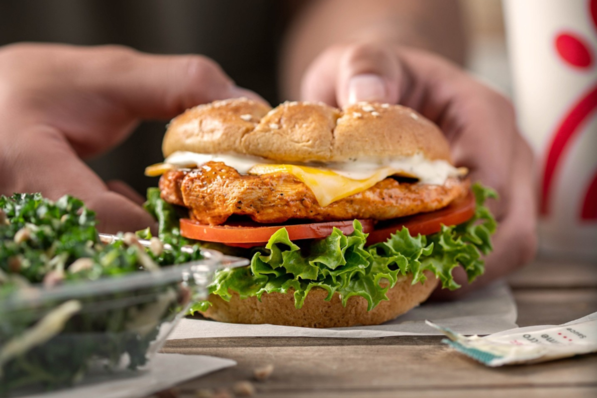 Chick-fil-A grilled chicken sandwich with lettuce, tomato, cheese, and sauce on a wheat bun, served with a side salad and a drink cup in the background.