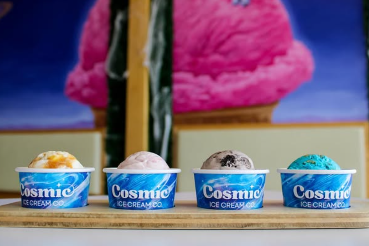 Four colorful scoops of Cosmic Ice Cream in blue branded cups, lined up on a wooden board with a bright pink ice cream mural in the background.