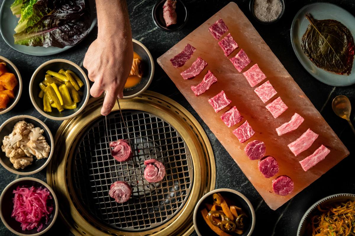 Korean BBQ setup with marbled raw beef slices on a salt block, assorted banchan sides, and a hand grilling meat over a tabletop grill on dark surface.