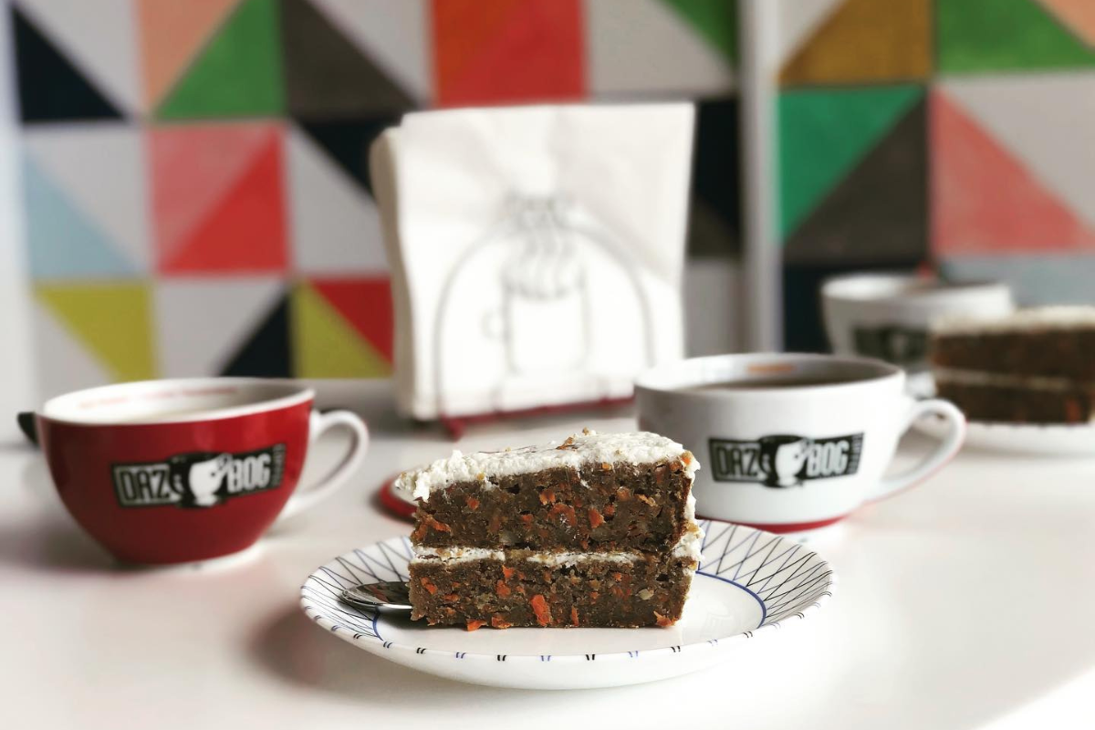 CL-Guacamole - DiningOut A slice of carrot cake with white frosting on a plate, served with Dazbog coffee in branded mugs, set against a colorful geometric wall.