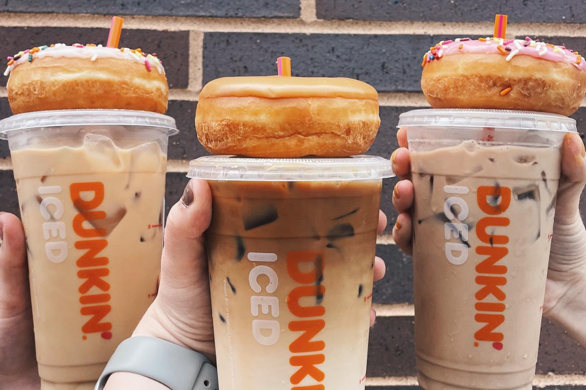 2_26_25_MONARCH_SMARIE-208 - DiningOut Three hands holding Dunkin' iced coffee drinks with donuts placed on top of each lid, two with sprinkles and one glazed, against a brick wall background.