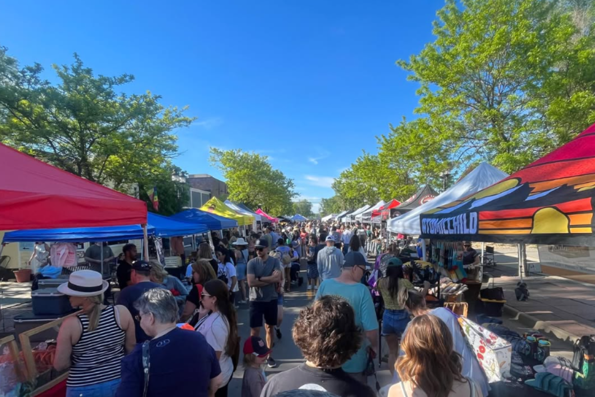 Shoppers walk through Erie Farmers Market on a sunny day, surrounded by colorful tents, green trees, and vendors selling handmade goods, produce, and treats.