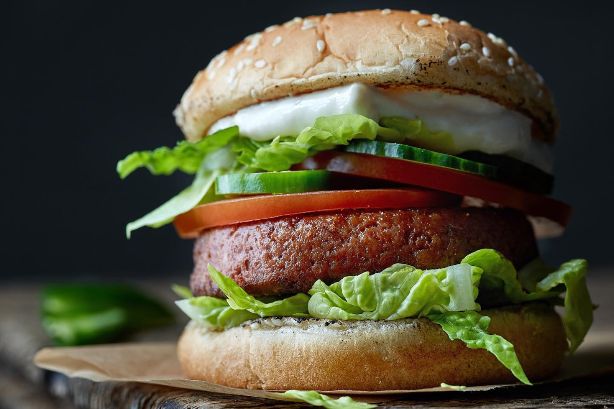 A close-up of a veggie burger stacked with lettuce, tomato, cucumber, and a sesame seed bun, served on parchment paper with a dark background.