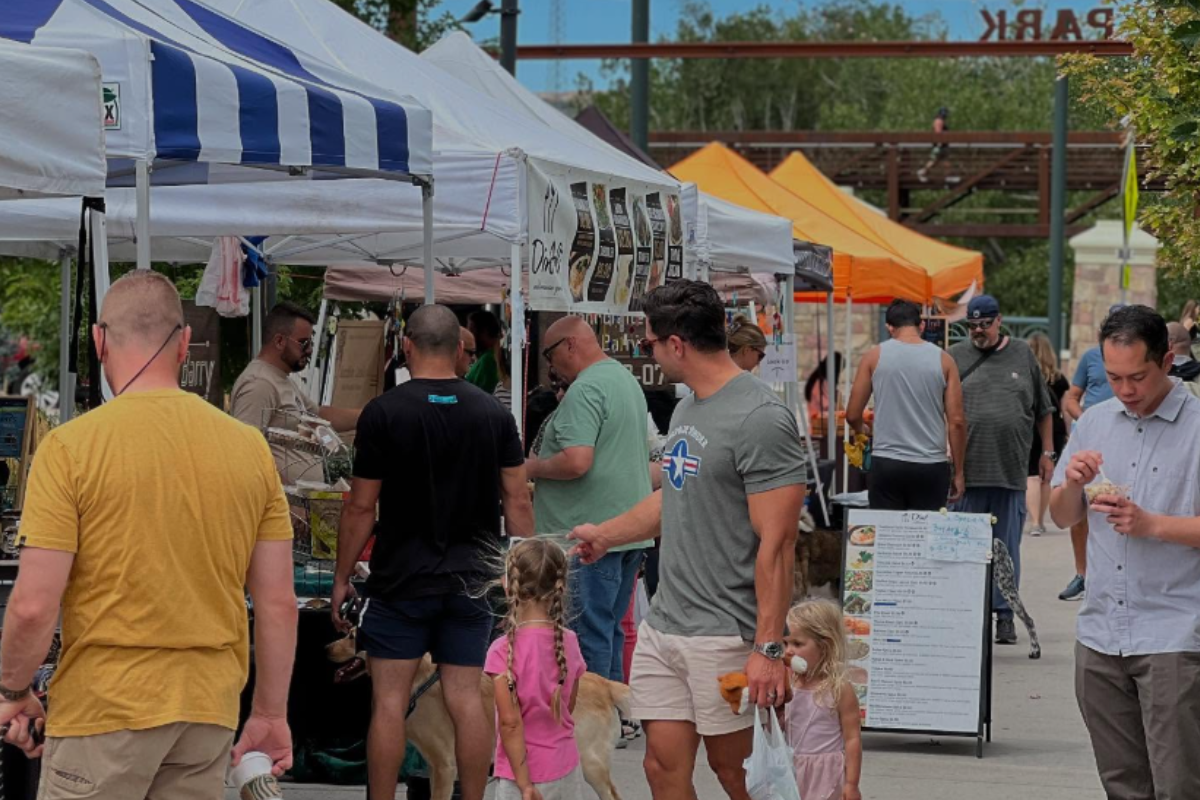 People walk with kids and shop at Festival Park Farmers Market in Castle Rock, surrounded by vendor tents with food, drinks, and handmade goods on display.
