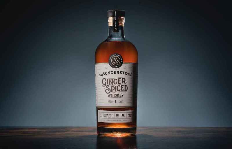 - DiningOut Bottle of flavored spirit labeled Misunderstood Ginger Spiced Whiskey against a slate gray background.