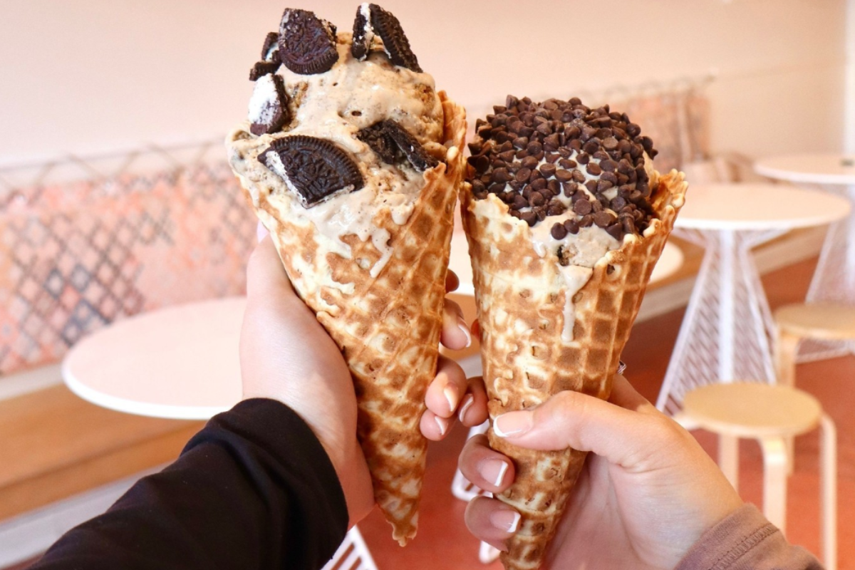Two waffle cones filled with Flower & Cream ice cream, one topped with Oreo cookies and the other with mini chocolate chips, held up inside a cozy ice cream shop.