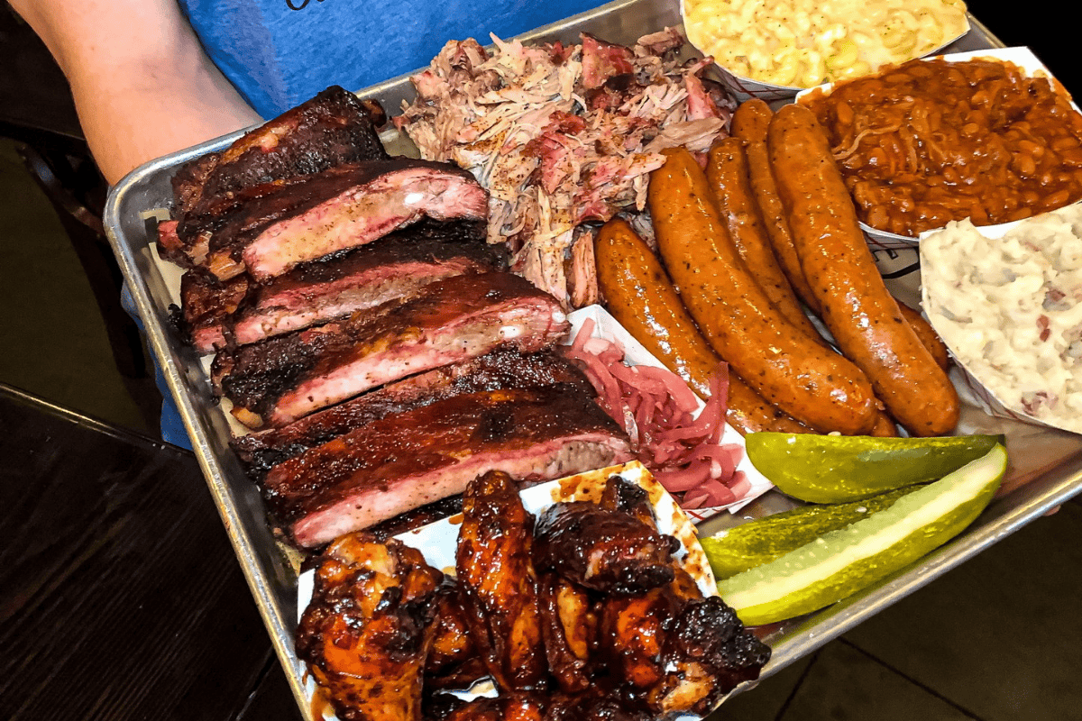 Tray from G-Que BBQ loaded with ribs, pulled pork, sausages, wings, mac and cheese, baked beans, potato salad, pickles, and pickled onions.