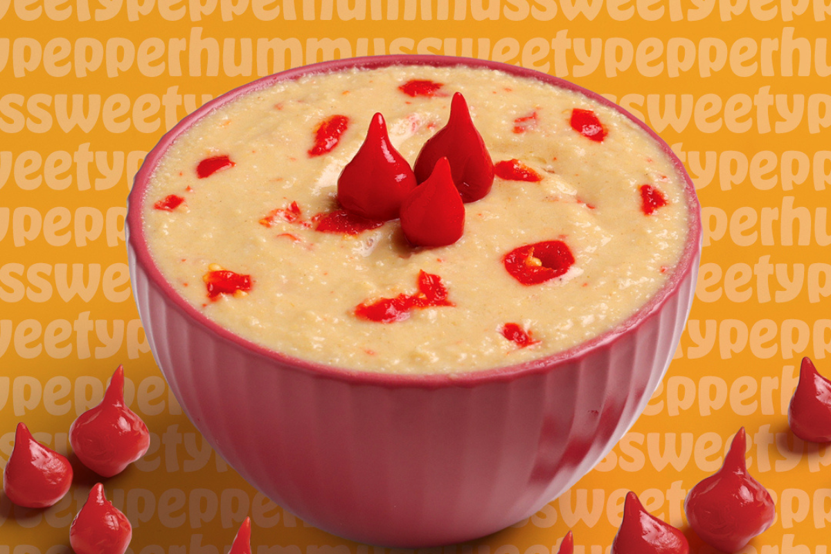 A pink bowl of sweet pepper hummus topped with red pepper drops, set against a bold orange background with playful repeating text.