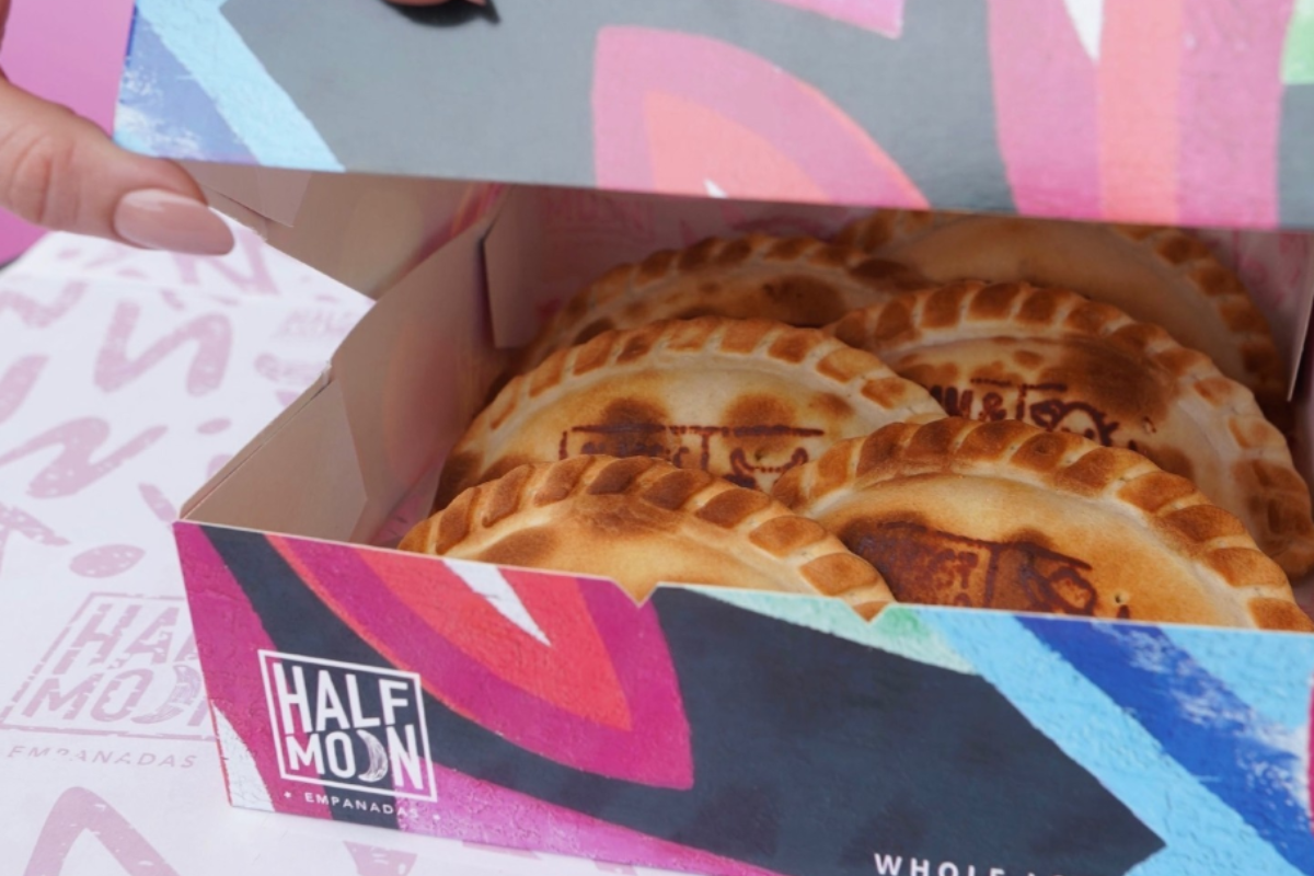 A close-up of a colorful box filled with golden-brown Half Moon empanadas, each stamped with a logo and packed neatly inside.