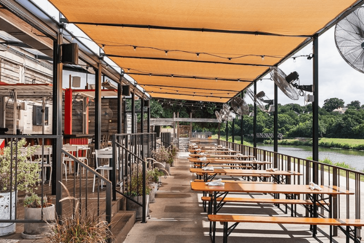 - DiningOut Outdoor patio at HG Sply Co. with shaded wooden picnic tables, ceiling fans, and views of green trees and a river in the background.