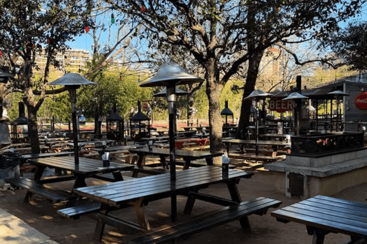 - DiningOut Outdoor beer garden with wooden picnic tables, heaters, string lights, and trees, offering a casual open-air space for dining and drinks.