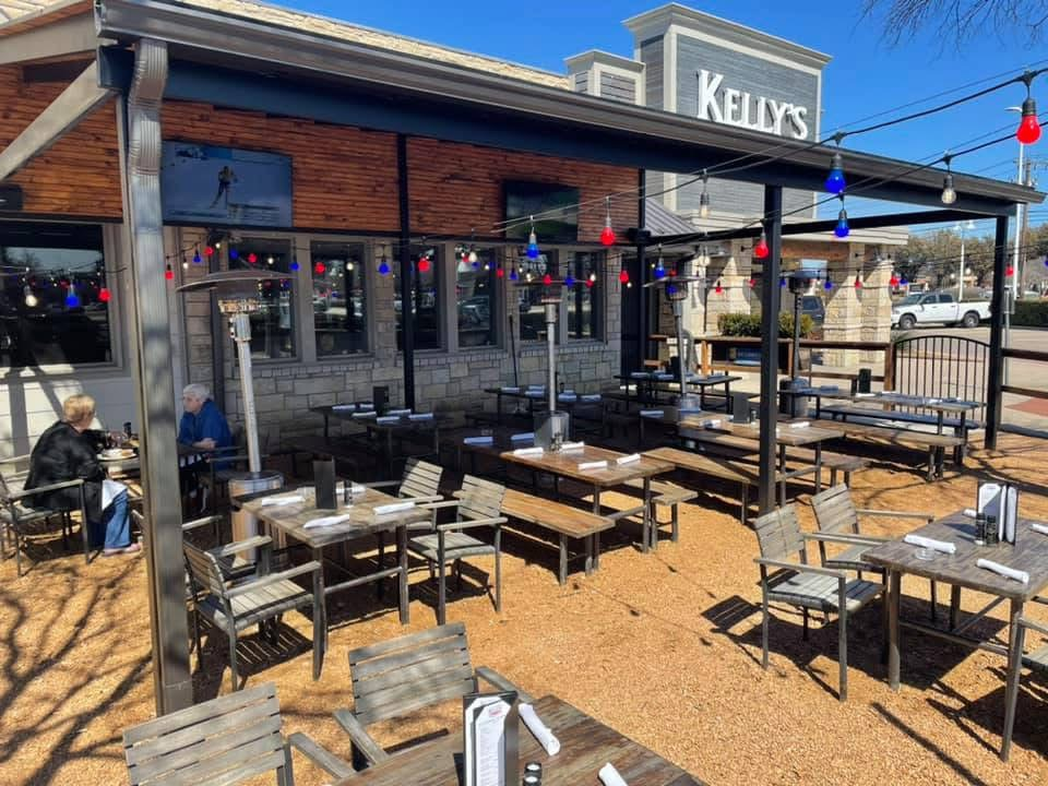 - DiningOut Outdoor patio at Kelly's Craft Tavern with wooden tables, chairs, string lights, and two people dining under a clear blue sky in The Colony.
