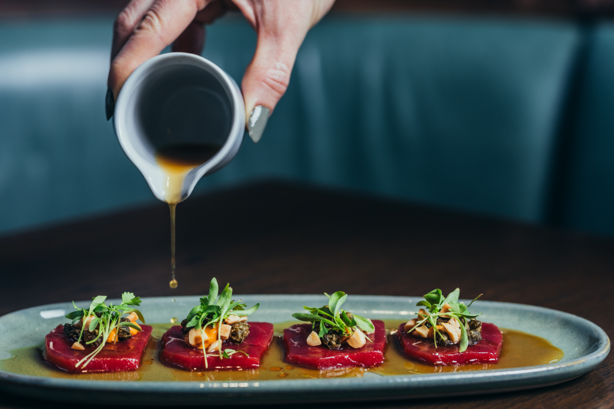 Hand pouring sauce over plated klaw tuna tataki topped with microgreens, caviar, and nuts on a green oval plate, set on a dark wood table.
