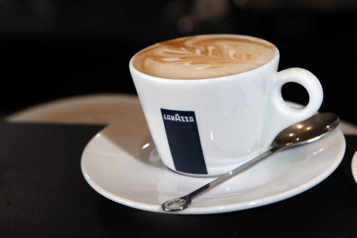 Where to Get the Best Matcha in Dallas Right Now - DiningOut A Lavazza-branded white cup filled with latte art on top, placed on a matching saucer with a silver spoon, set against a dark background.