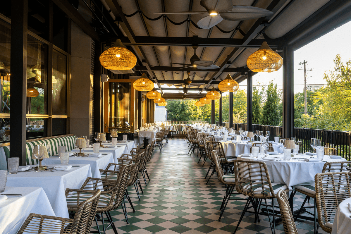 - DiningOut Outdoor patio at Le Passage with elegant tables, white linens, wicker chairs, hanging woven lights, ceiling fans, and a view of trees in the background.