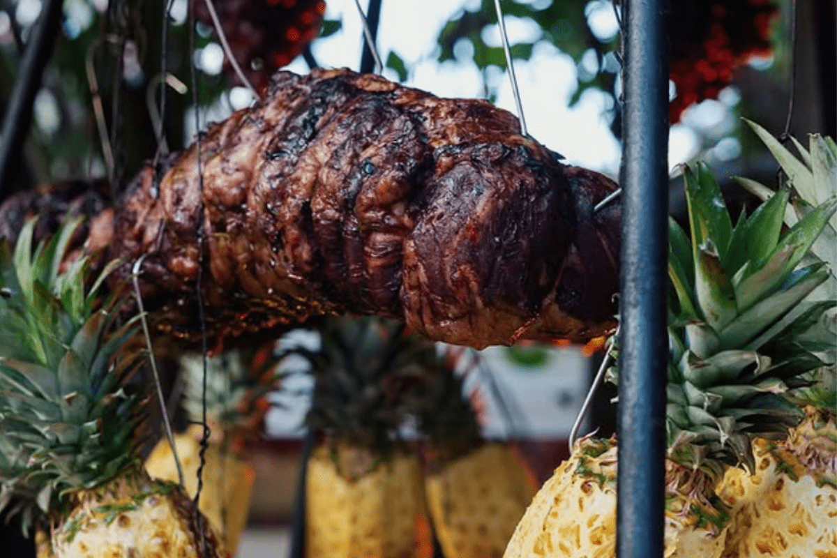 Grilled meat roasting over open fire, surrounded by hanging pineapples at Los Fuegos by Francis Mallmann, showcasing open-flame cooking style.