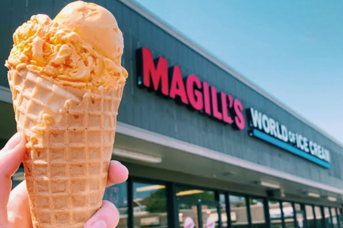 Tipping-your-server - DiningOut Close-up of an orange ice cream cone held in front of a storefront with a bright sky, showing creamy texture and crunchy waffle cone details.