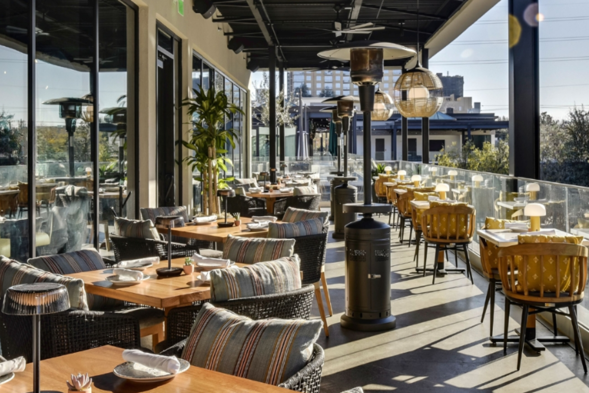 - DiningOut Outdoor patio at Mexican Sugar with wooden tables, modern wicker and cushioned chairs, hanging lights, patio heaters, and glass railing with city views.