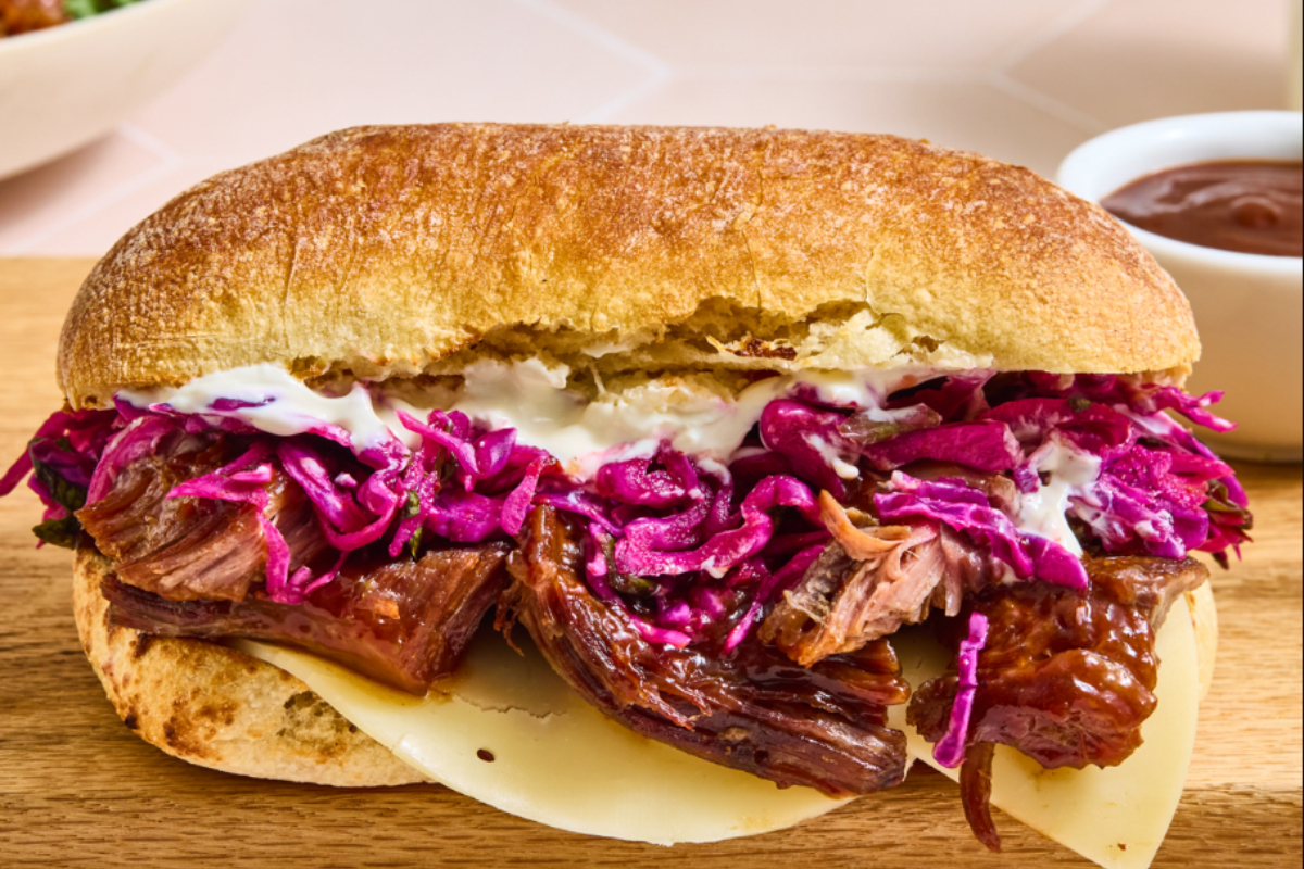 Close-up of a pulled beef sandwich with melted cheese, creamy sauce, and vibrant purple slaw on toasted ciabatta bread, served with a side of BBQ sauce.