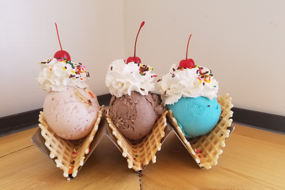 spaghetti-bolognese - DiningOut Three waffle cone bowls filled with colorful ice cream scoops, topped with whipped cream, rainbow sprinkles, and cherries, on a wooden table.