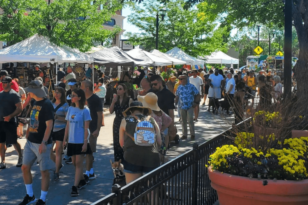 Shoppers walk through the busy Parker Farmers Market on a sunny day, browsing vendor tents filled with food, flowers, and handmade goods under shady trees.