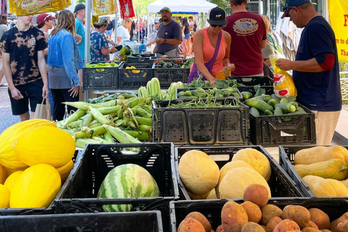 People + Produce at Belleview Station in Denver with shoppers browsing fresh produce like melons, corn, cucumbers, and potatoes under shaded vendor tents.