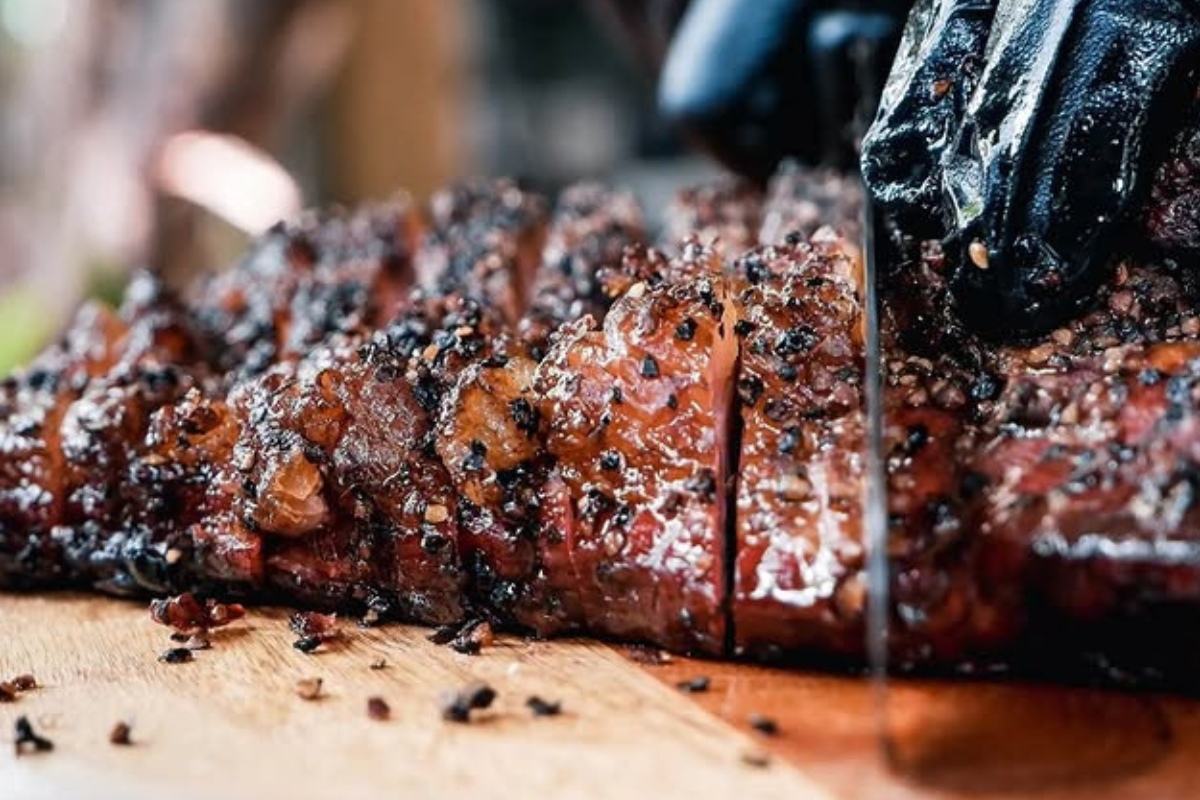 Close-up of sliced brisket from Piggin’ Out Smokehouse, glistening with fat and cracked pepper, being cut on a wooden board by a gloved hand.

