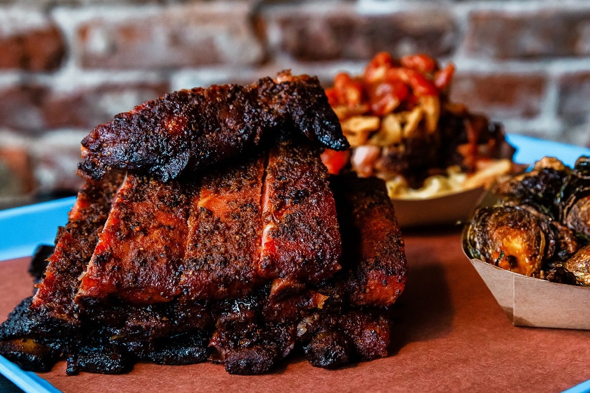 Stack of smoked pork ribs with a dark bark and red smoke ring, served on a tray with crispy Brussels sprouts and loaded fries in the background.