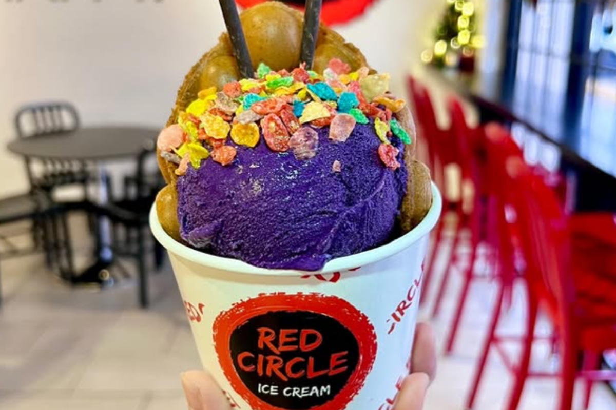 Cup of Red Circle Ice Cream with vibrant purple scoop, fruity cereal toppings, and bubble waffle, held in a shop with red chairs and blurred background.