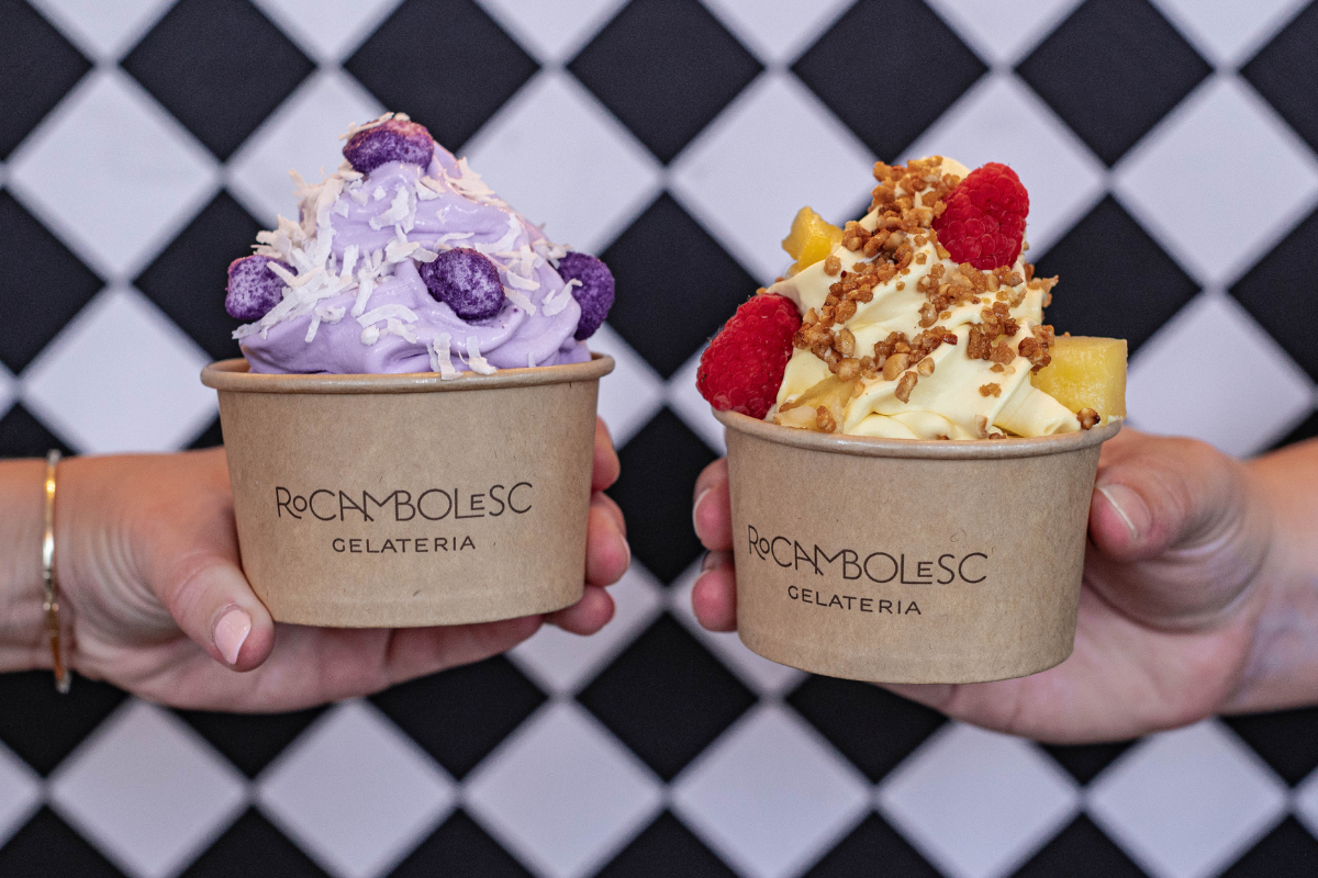 Two hands holding cups of Rocambolesc gelato, one topped with coconut and purple candies, the other with raspberries, mango chunks, and crumble, against a tiled wall.