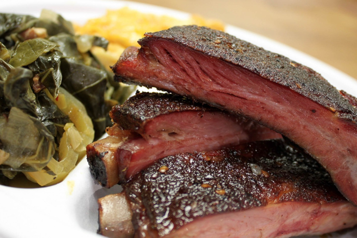 Close-up of smoked pork ribs from Rolling Smoke BBQ, served with tender collard greens and creamy mac and cheese on a white plate.

