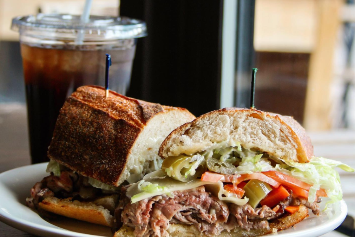 Salt & Grinder sandwich stacked with roast beef, shredded lettuce, tomatoes, peppers, and cheese on crusty bread, served with iced soda in the background.