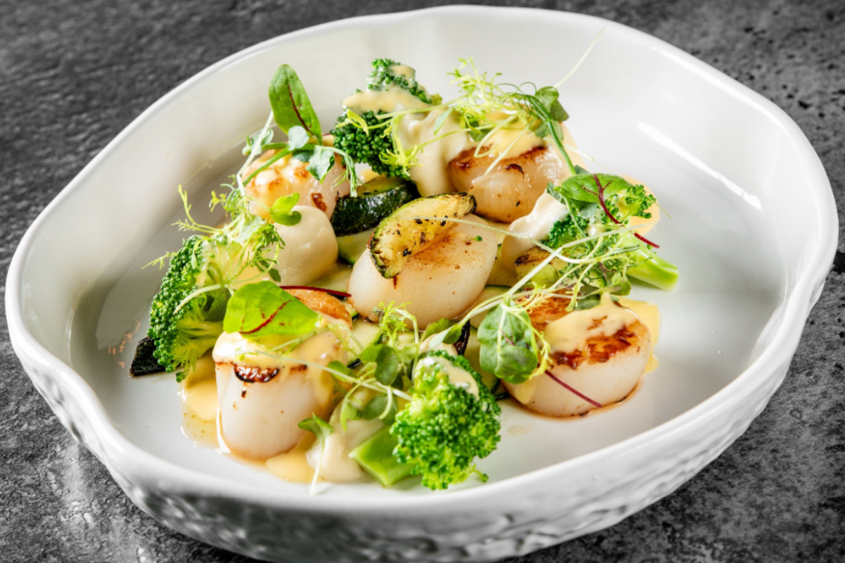- DiningOut Seared scallops served with broccoli, zucchini, and microgreens, topped with a creamy sauce in a white bowl on a dark stone surface.