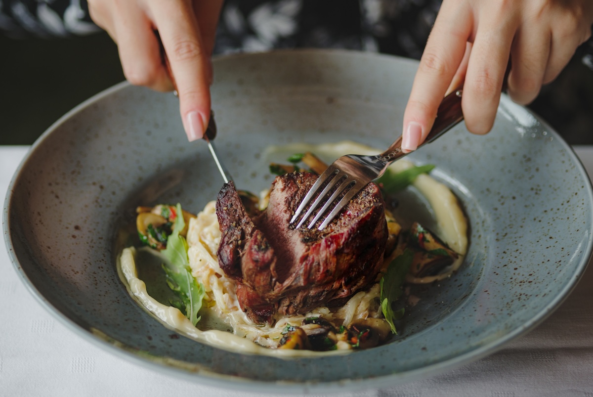 Two hands are cutting a piece of beef in a plate. A close-up picture of a blue round plate full of tasty dish on a white table background. Delicious expensive food. Restaurant, food concept.