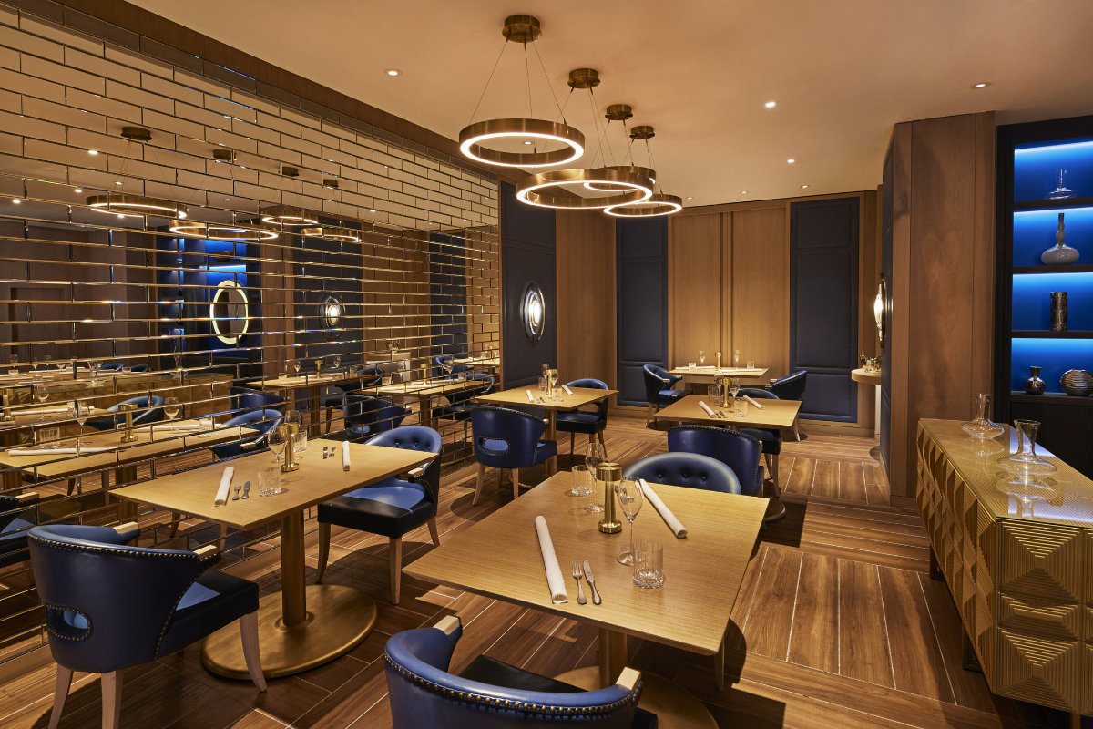 Elegant modern restaurant with wood floors, navy blue chairs, gold accents, circular pendant lights, and mirrored brick wall creating a stylish dining space.

