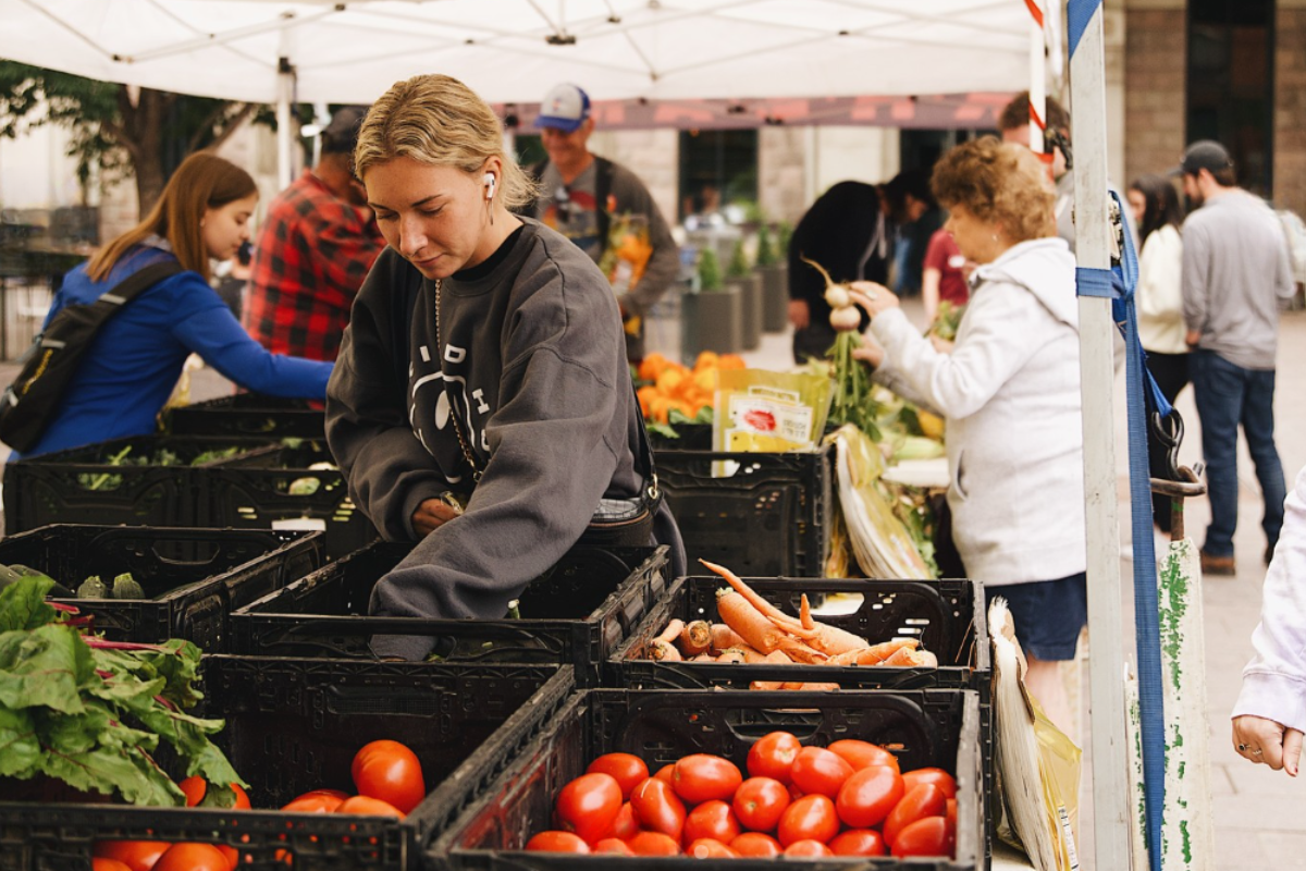 Shoppers at Union Station Urban Market in downtown Denver browse crates filled with fresh tomatoes, carrots, and leafy greens under a white canopy tent.