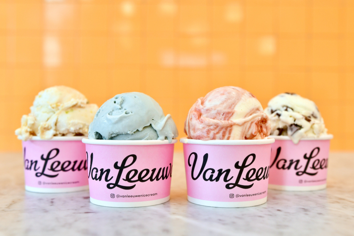 Four colorful Van Leuwen ice cream scoops served in pink cups on a marble counter, with a bright orange tiled wall in the background.