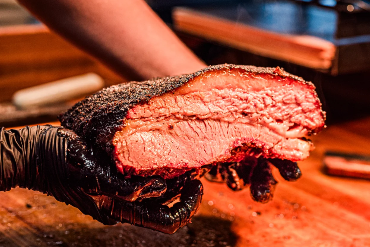 Close-up of a juicy smoked brisket slice from Wayne's Smoke Shack, held in gloved hands, showing its bark, fat cap, and pink smoke ring.