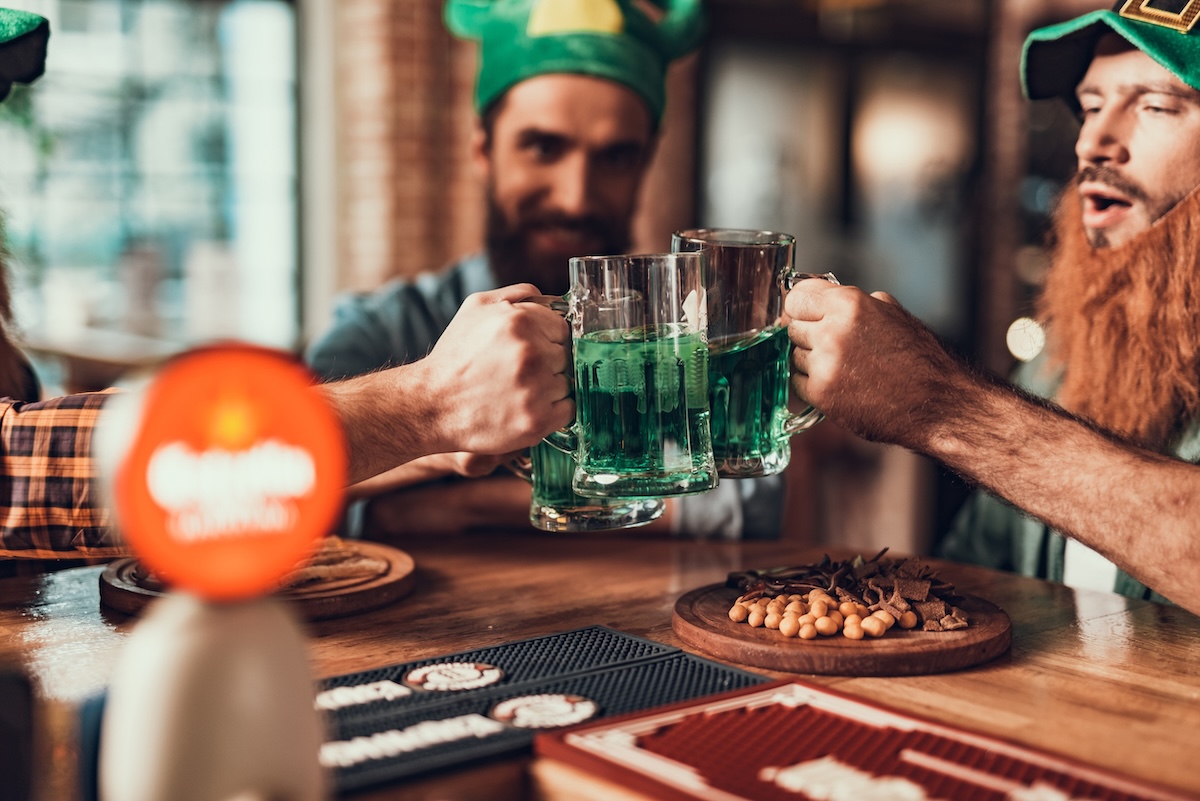 Close up of male hands clinking mugs of traditional green beer. Gentlemen celebrating Saint Patrick Day while sitting at bar counter with snacks by_iakovenko ID_114840534