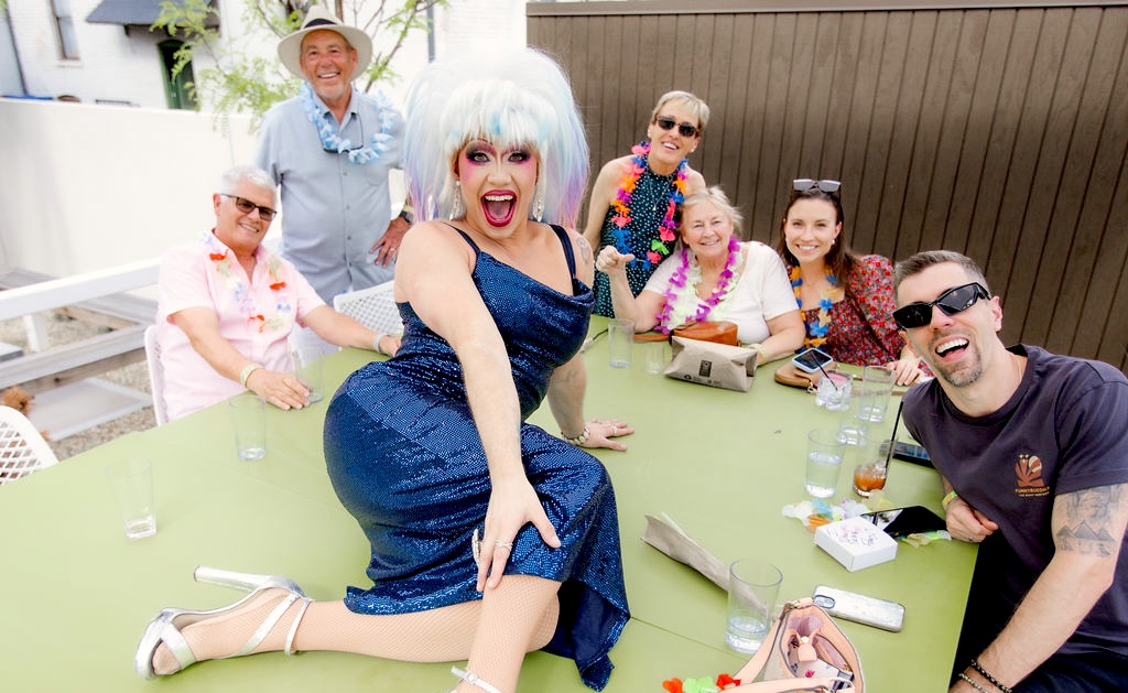 Champagne Tiger_Anastasia Krystals - DiningOut Drag queen in a sparkling blue dress poses on a green outdoor table, surrounded by laughing friends at a backyard party.