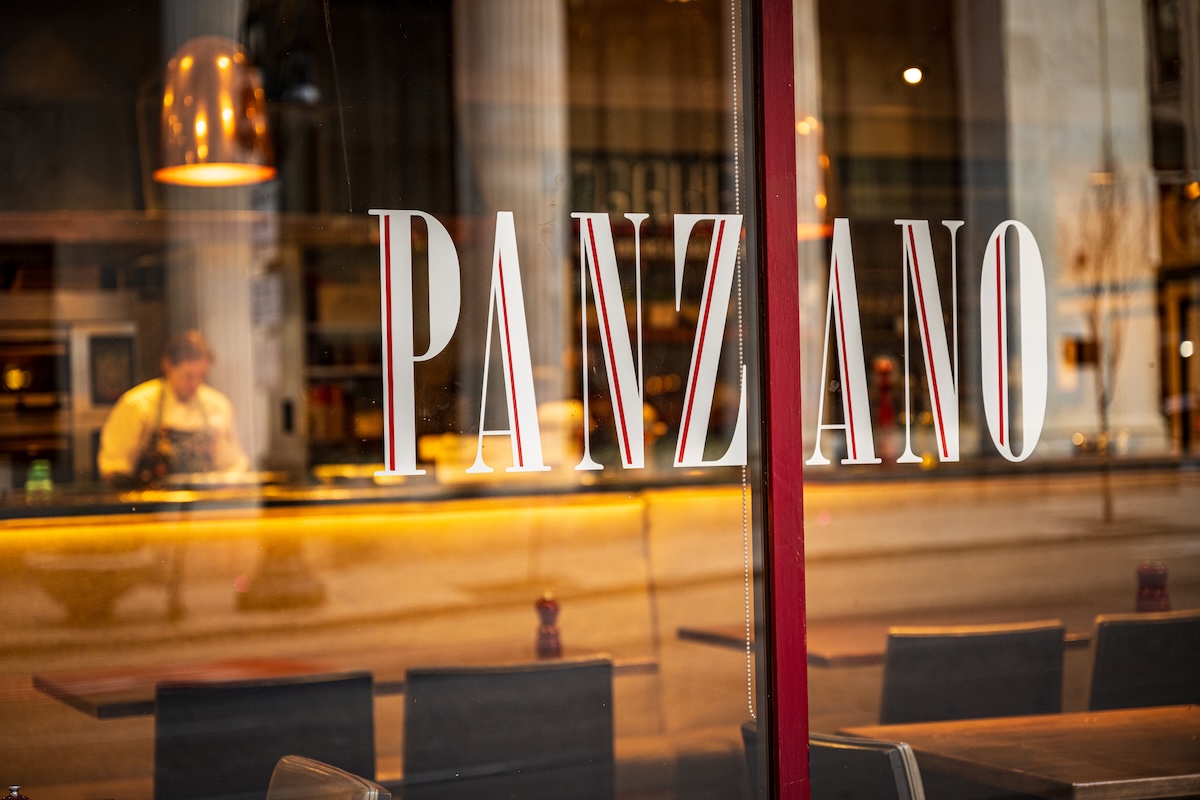Restaurant window with the name 'PANZANO' in tall white letters; warm interior light and a chef working in the background.