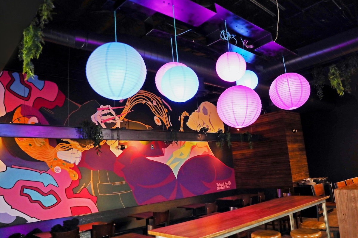 Colorful mural walls with abstract shapes, hovering blue and pink paper lanterns over a long wooden table and stools in a dimly lit cafe/bar area
