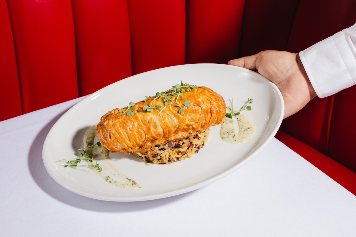 Golden flaky pastry sandwich resting on a bed of herbed rice, garnished with microgreens on a white oval plate.