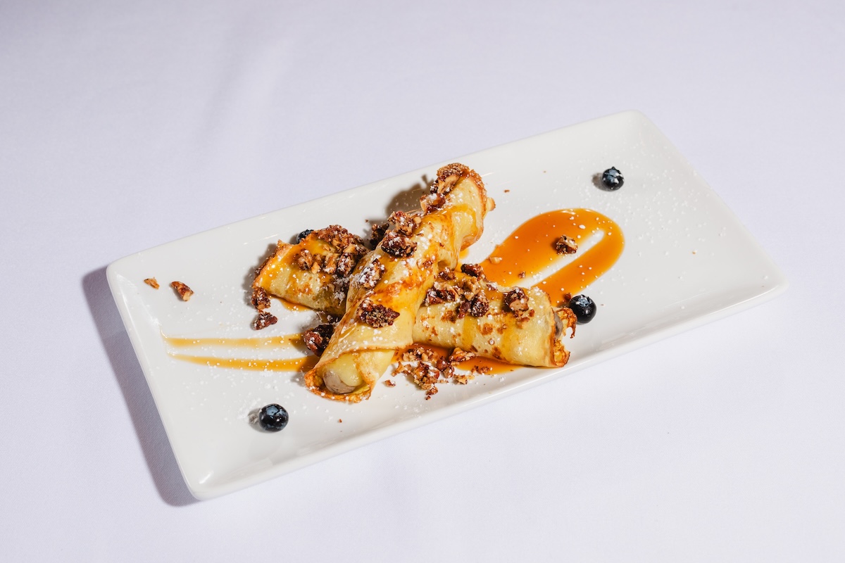 Two rolled crepes topped with caramel sauce and crushed nuts on a white rectangular plate, with blueberries garnish.