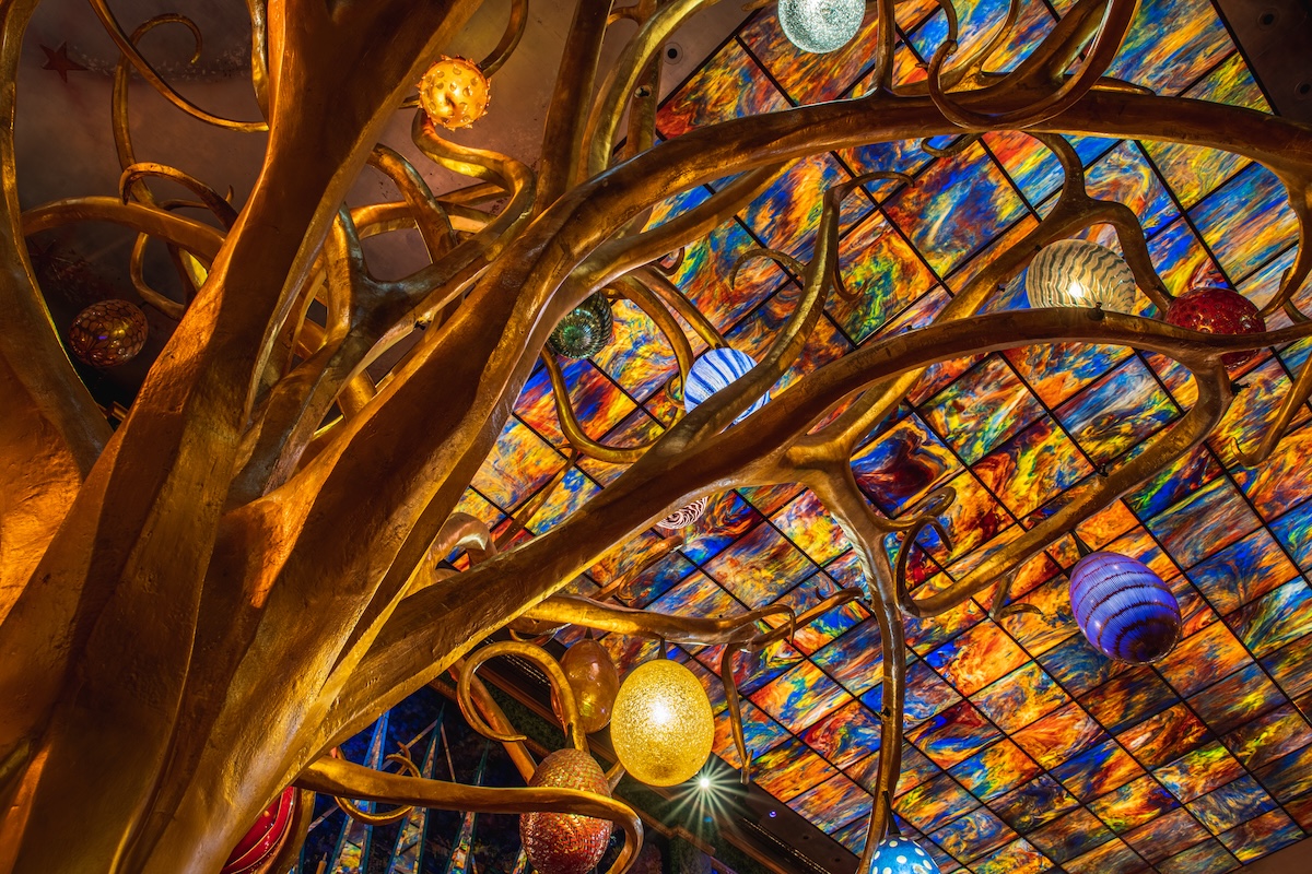 Golden tree sculpture with twisting branches beneath a vibrant multicolored glass ceiling, hanging glass ornaments.